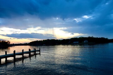 Obraz premium Dramatic Sunset Storm Clouds Over Peaceful River with a Wooden Pier and Reflections