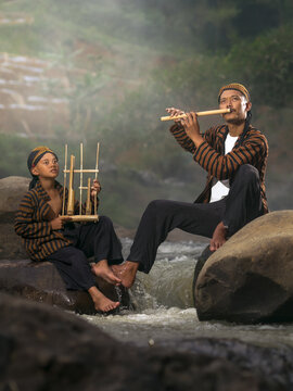 Traditional Sundanese musicians playing angklung and bamboo flute by a river in rural Indonesia.