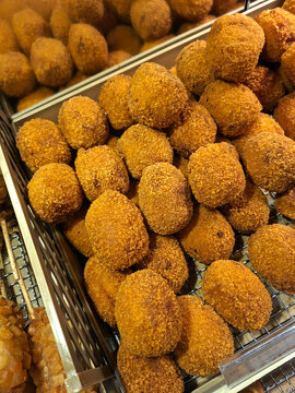 A close-up shot of numerous golden-brown fried potato croquettes or cheese balls piled high on a metal cooling rack inside a display case