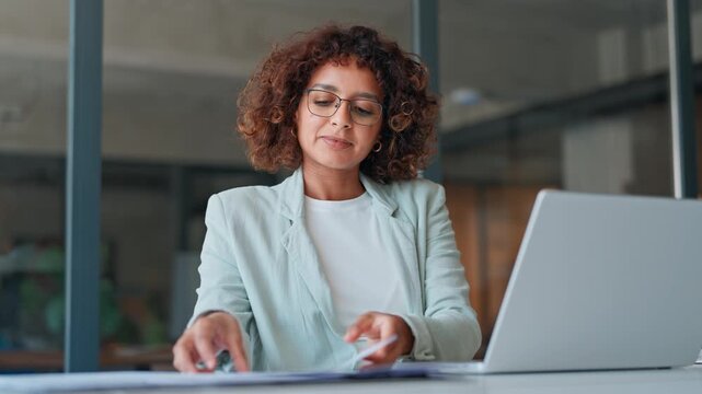 Young curly accountant entrepreneur manager businesswoman doing paperwork using pc. Focused latin hispanic business woman working on laptop computer reading financial tax document report in office.