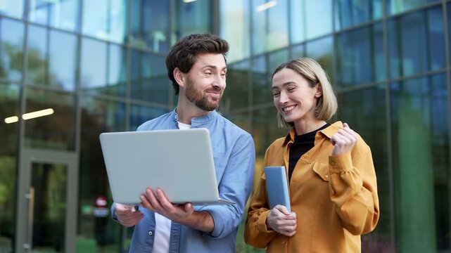 Happy business colleagues react to good news on a laptop near a modern office building. Businessman and businesswoman smile after reading positive information and share excitement outdoors. Close up
