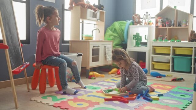Girl sits on orange stool and plays with wooden blocks on foam number mat in building; quiet concentration and childhood learning.