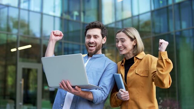 Close-up of happy business colleagues viewing successful results on a laptop near a modern office building. Businessman and businesswoman celebrate a profitable deal and company growth outdoors.