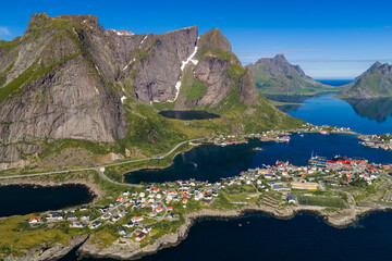 Aerial view of Lofoten village near Reine and Reinebringen, Norway. Colorful houses and red rorbuer line fjords and lakes, summer light reveals snow, roads, and glassy water. © Aerial Film Studio