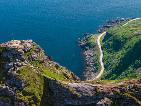 Aerial view shows a rugged cliff above turquoise waters, a serpentine coastal road, and a narrow hiking path in Norway's Lofoten Islands under summer sun.