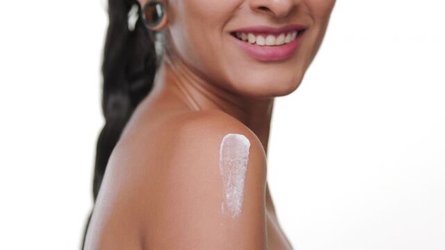 Big close-up shot of latin woman with bronzed skin applying nourishing cream on shoulder on studio white background | Skin care advertisement