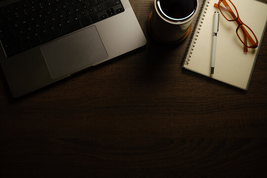 Dark wooden desk workspace with laptop, coffee cup, notebook, pen, and eyeglasses, ideal for remote work and productivity concepts.