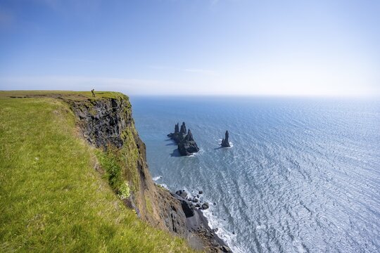 Young man standing on cliff, view from steep cliff to the rocks Reynisdrangar in the water, at Reynisfjara beach, Vik, South Iceland, Iceland