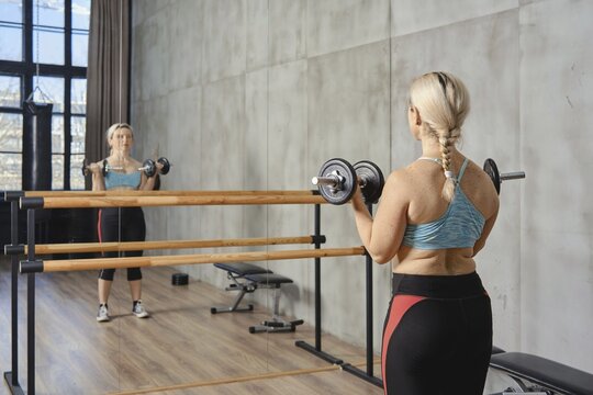 A woman with a braid exercises with dumbbells in a contemporary gym. She is focused, using a mirror for form checks. Natural light fills the space, enhancing the atmosphere
