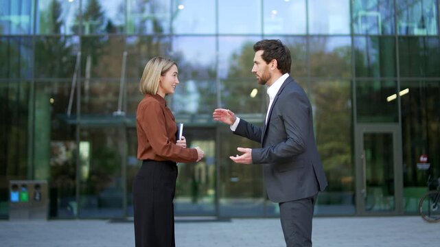 Businessman and businesswoman greet each other with a handshake outside a modern office building. Following the introduction, he invites her to continue meeting inside, proceed together in discussion.