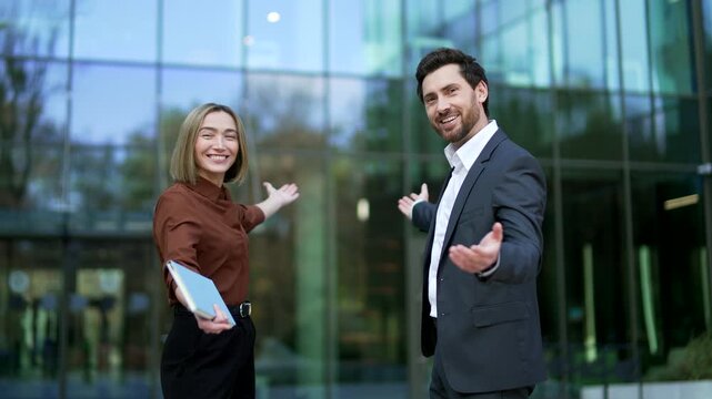 Businessman and businesswoman stand near entrance of office building, looking at camera and inviting viewers to join their team. Friendly professionals promote hiring, partnership, new opportunities.