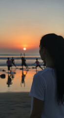 Fototapeta premium Woman watches a group of people practicing yoga on the beach during a peaceful golden sunset.