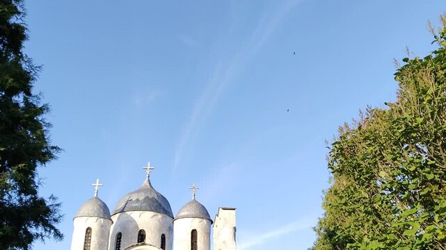 Cinematic panning shot of ancient Russian Orthodox cathedral of Ioann Predtecha (John the Precursor) dome peaks in Pskov shifting toward clear blue sky and lush green trees before smoothly returning