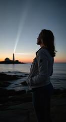 Fototapeta premium Woman watches a lighthouse beam cutting through the dusk sky while standing on a coastal rock formation.