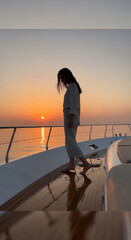 Fototapeta premium Woman walks along the wooden deck of a yacht at sunset while the golden light reflects off the calm water surface.