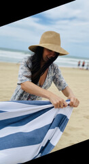 Fototapeta premium Woman wearing a sun hat unfolds a blue and white striped beach towel while standing on the sandy shore.