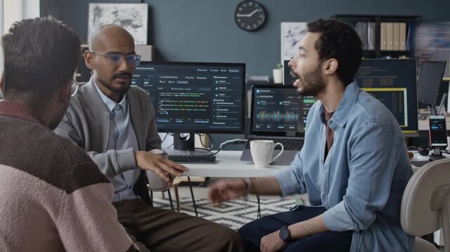 Handheld shot of multiethnic young male programmers discussing software development project during coffee break in office with running codes on screens. Suitable for tech startup marketing