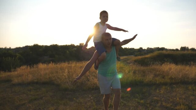 Happy father and son playing on the field at sunset. Parent with kid having fun together outdoor. Little boy sitting on shoulders of his daddy and playing raised hands as airplane. Slow motion