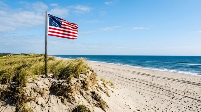 The flag of the united states of america waving on a pole atop sandy dunes with tall beach grass, near the blue ocean water on a clear sunny day