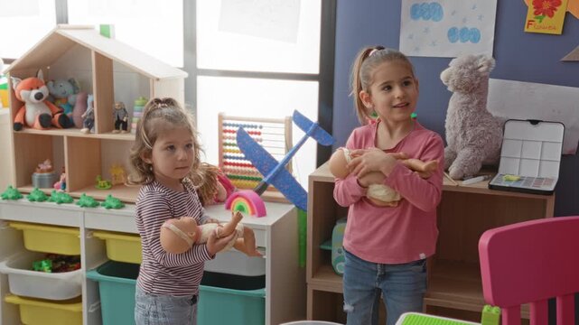 Two girls cradling baby dolls and smiling while standing by a toy shelf and abacus in a preschool playroom building; joyful play.