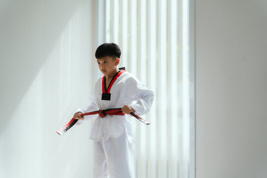 Young boy practices martial arts in a bright room during the day while wearing a taekwondo uniform and holding a black belt