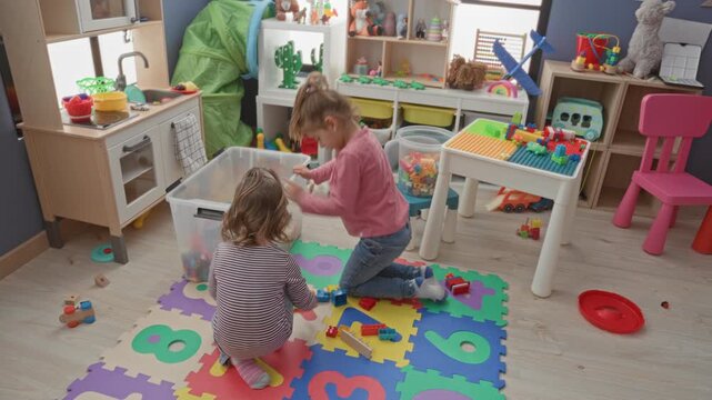 Girl kneeling by clear toy bin, reaching for colorful blocks and playing on foam numbers mat inside a playroom building; playful childhood bonding.