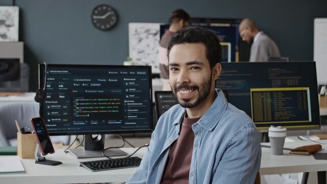 Chest up slowmo portrait of Middle Eastern young man sitting at computer desk with running codes posing for camera holding coffe cup and smiling. Ideal for tech recruitment and software development ad
