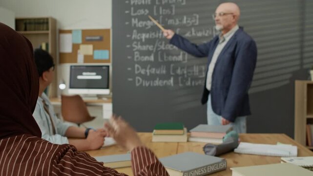 Young Muslim student in hijab raising hand during business English lesson with senior Caucasian teacher pointing at blackboard. Clip is perfect for promoting language schools and diversity