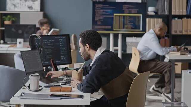 Medium side view shot of young Middle Eastern male employee eating noodles from takeout box while coding on laptop in IT office. Ideal for corporate lunch break, software developer lifestyle campaigns