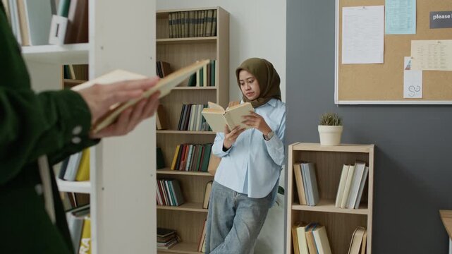 Young adult Asian Muslim female student wearing hijab standing in library, reading book while classmate holds book in foreground. Clip is ideal for marketing campaigns promoting inclusive education