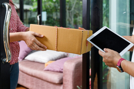 Close up of delivery man handing cardboard parcel box to customer at home doorway. Delivery person holding tablet for signature while customer receiving package at door.