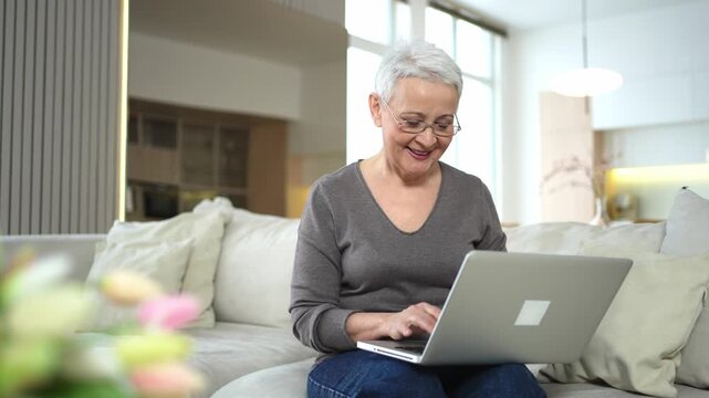 Confident stylish happy middle aged senior woman using laptop at home. Stylish older mature 60s grandmother sitting at couch looking at computer screen typing chatting reading writing email