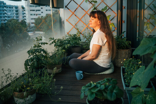 Woman enjoying peaceful sunrise moments while sitting among plants on a balcony garden in the city. Mindfulness, mental wellbeing, slow living and connection with nature