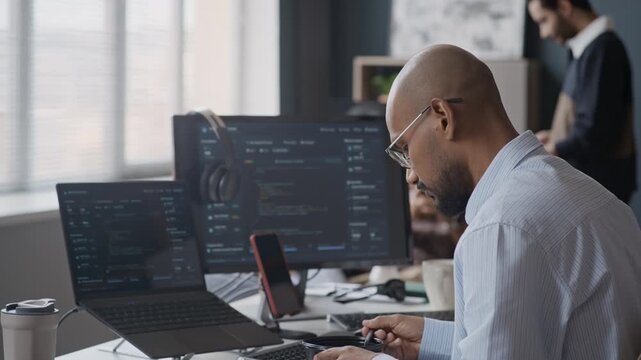 Side view shot of young Indian male software developer eating lunch at desk while looking at computer screens with code in IT office. Footage suits commercials for tech companies and office life
