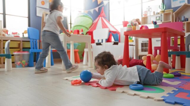 Little hispanic girl crawling on foam mat reaching for a toy beside colorful table and chalkboard inside a childcare building; playful curiosity.