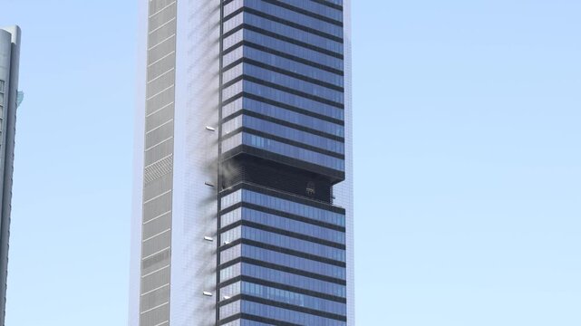 Dangerous fire burning on an upper floor of a modern office skyscraper against a clear blue sky