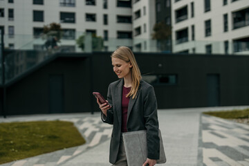 Fototapeta premium Young businesswoman smiling using smartphone walking city street
