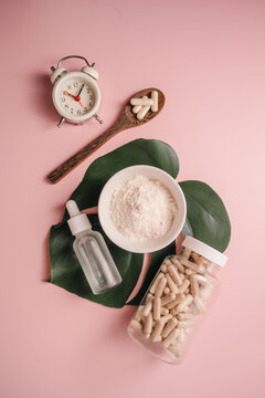 Collagen powder in a bowl with food supplement capsules, on monstera leaf with alarm clock on pink background, Healthy aging concept