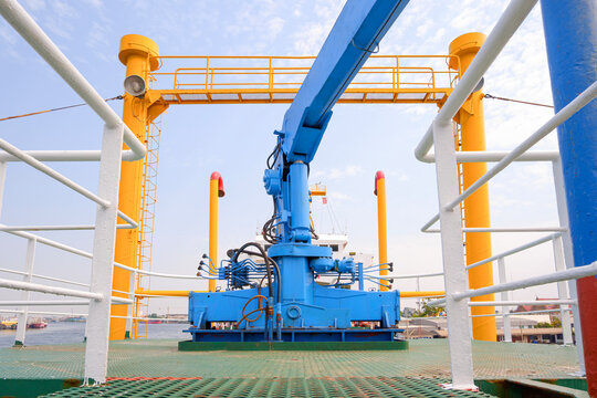 Blue hydraulic marine crane mounted on an industrial ship deck. Features safety yellow and white railings against cloudy sky. Ideal for maritime industry and technical content