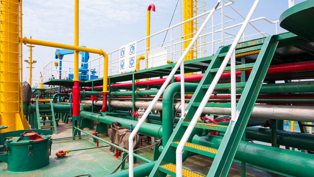 Colorful industrial oil tanker ship deck with complex pipes, green walkways, and white ladders. Features safety signs under a partly cloudy sky.