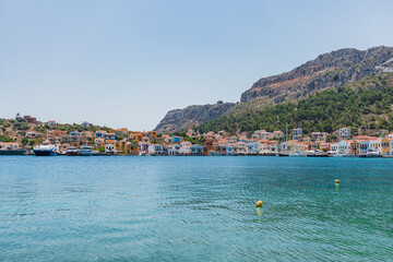 Fototapeta premium Coastal view of Kastellorizo, Megisti, Greece. Colorful buildings line the waterfront, surrounded by clear blue water and green hills in the background.
