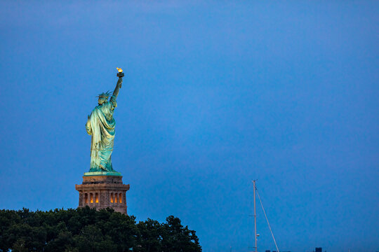 New York, New York, United States - 08 July 2021: View of the Statue of Liberty illuminated against a deep blue twilight sky, with trees at the base and a sailboat mast.