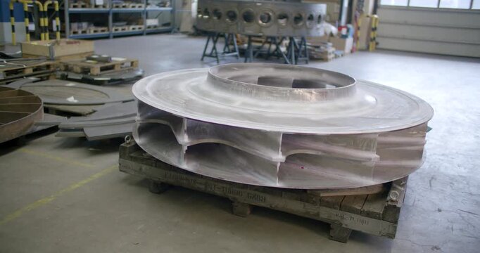 A large circular metallic fan or turbine component resting on a pallet in the middle of an industrial manufacturing plant