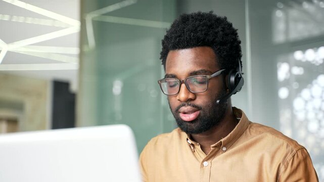 Close up of focused African American customer support agent wearing headset and talking. Professional black man operator working on helpline telesales via laptop inside corporate business office.