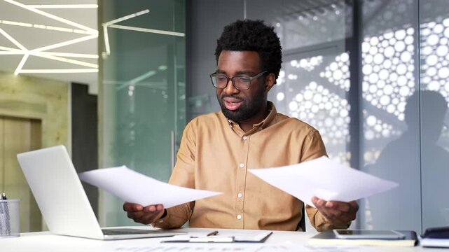 Confused African American businessman faces difficulty with complex paperwork. Frustrated employee financier looks at documents inside corporate office workplace and cannot understand problem.