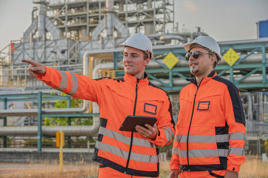 Industrial engineers in safety uniforms reviewing blueprint at oil refinery plant, teamwork inspection concept, heavy industry maintenance, energy production site