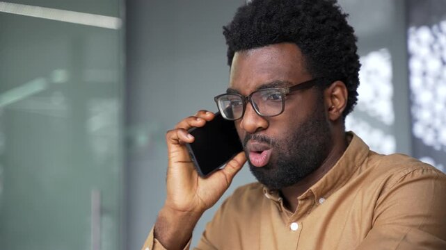 Close up of angry African American boss shouting and arguing during smartphone call. Furious black man executive manager talking aggressively and scolding someone on mobile phone in office.