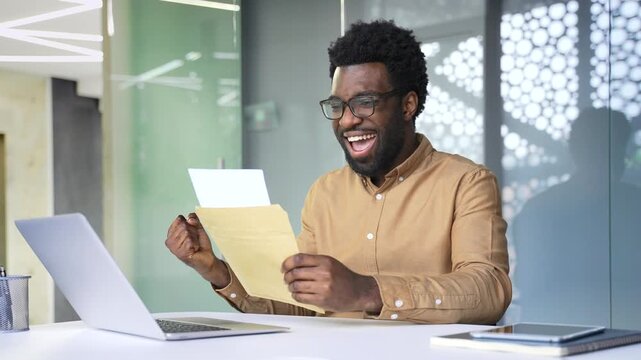 African American businessman reading letter with great news, feeling excited and celebrating success. Happy surprised black man manager sitting at workplace desk in office with genuine joy expression.