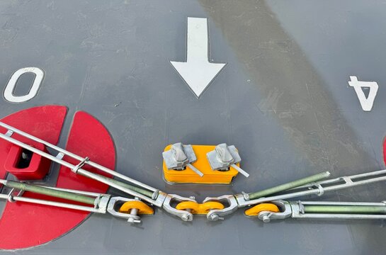 Описание): Close up of industrial steel turnbuckles, hooks and lashing tension chains securing cargo on a vessel deck with painted white arrow and red elements