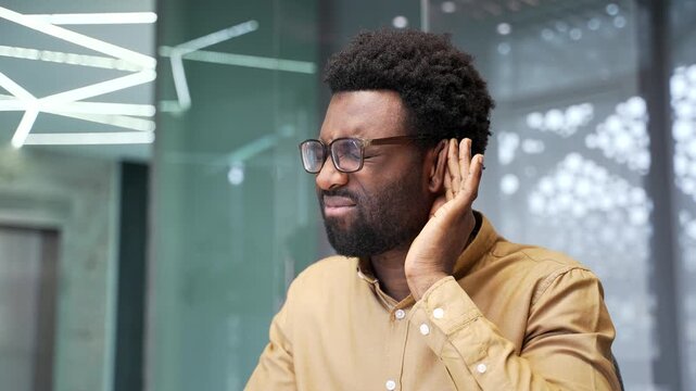 Close up of unwell African American businessman who has earache at work. Sick black man worker sitting at corporate office desk touches painful ear with fingers because of otitis media indoors.